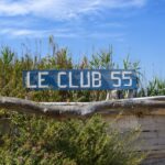 Rustic Le Club 55 entrance sign surrounded by coastal greenery under a clear blue sky in Saint-Tropez, France.