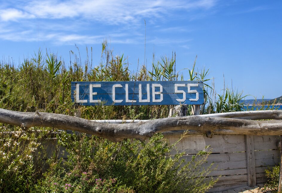 Rustic Le Club 55 entrance sign surrounded by coastal greenery under a clear blue sky in Saint-Tropez, France.