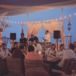 Guests dining at dusk under festoon lights at Verde Beach, with a live music setup and the sea behind