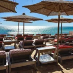 Sunbeds with red cushions under straw umbrellas beside the sea at Byblos Beach Ramatuelle