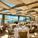 Round dining tables beneath a draped canopy with sea views at Byblos Beach Ramatuelle