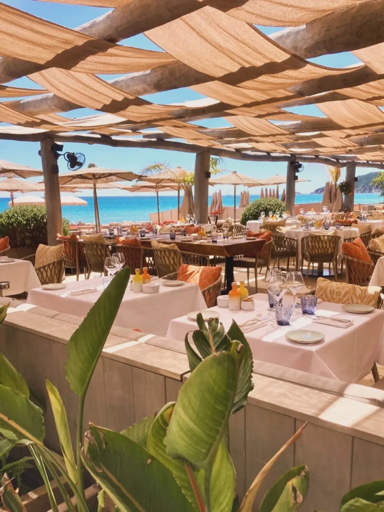 Restaurant tables framed by greenery with a bright sea view at Byblos Beach Ramatuelle