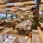 Dining terrace with white-clothed tables under a woven canopy at Byblos Beach Ramatuelle
