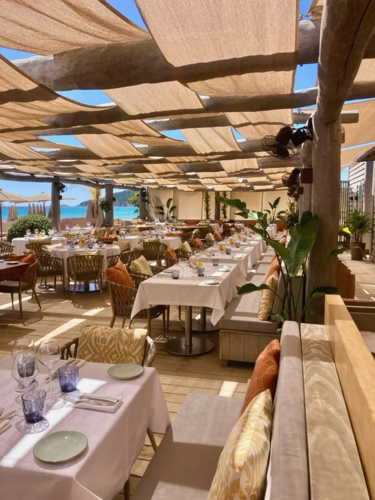 Dining terrace with white-clothed tables under a woven canopy at Byblos Beach Ramatuelle