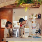 Staff working the counter with a beer tap beneath a hanging fern at El Caminito Palma
