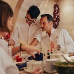 Two chefs conferring at the pass under a stone arch hung with dried peppers at El Caminito Palma