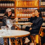 Two guests chatting over drinks at a table against the lit wine wall at El Caminito Palma