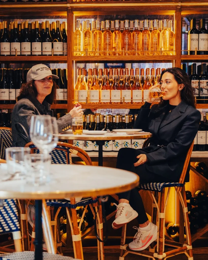 Two guests chatting over drinks at a table against the lit wine wall at El Caminito Palma
