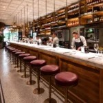 Chefs prepping behind the marble counter lined with burgundy stools and a backlit wine wall at El Camino Restaurant