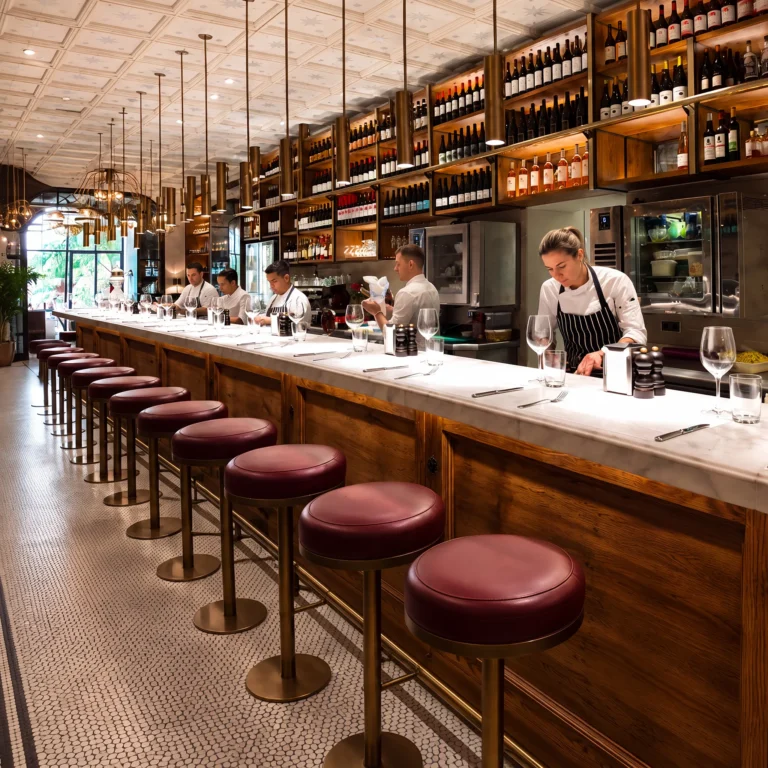 Chefs prepping behind the marble counter lined with burgundy stools and a backlit wine wall at El Camino Restaurant