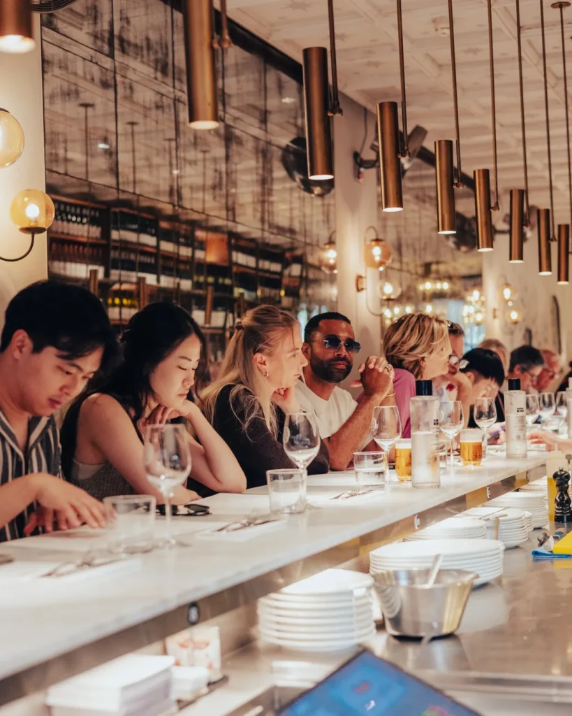 Guests with beers and wine seated along the marble counter under an antiqued mirror at El Camino Restaurant