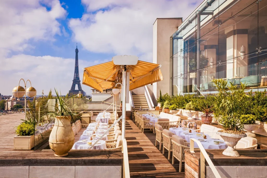 Rooftop terrace lined with set tables and a fringed umbrella facing the Eiffel Tower at Gigi Rigolatto Paris