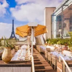 Rooftop terrace lined with set tables and a fringed umbrella facing the Eiffel Tower at Gigi Rigolatto Paris