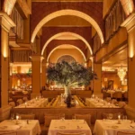 Vaulted brick-arched dining hall with a central olive tree and white-clothed tables at Gigi Rigolatto Paris