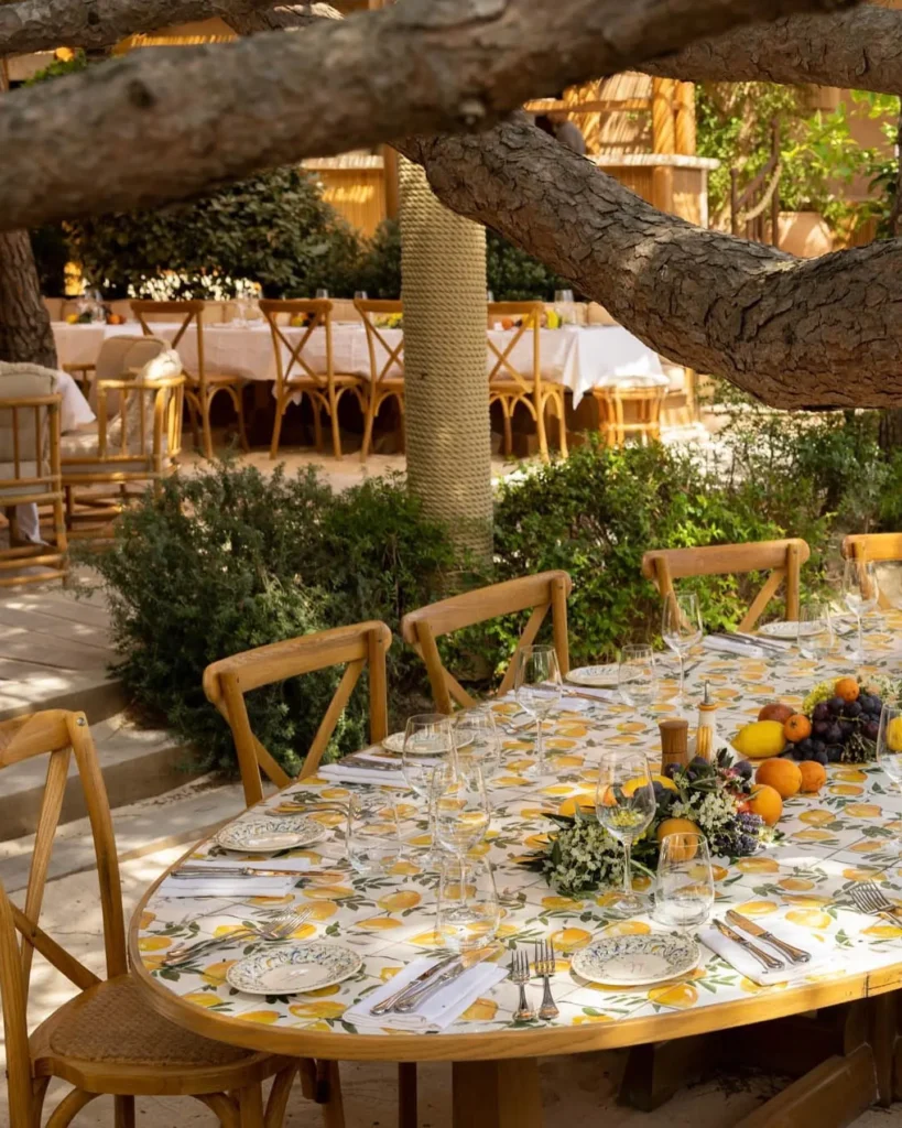 Oval table dressed in a lemon-print cloth with fruit and flowers under a pine at Gigi Rigolatto Saint-Tropez