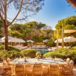 Set dining table on the sand with the pool and parasols beyond at Gigi Rigolatto Saint-Tropez