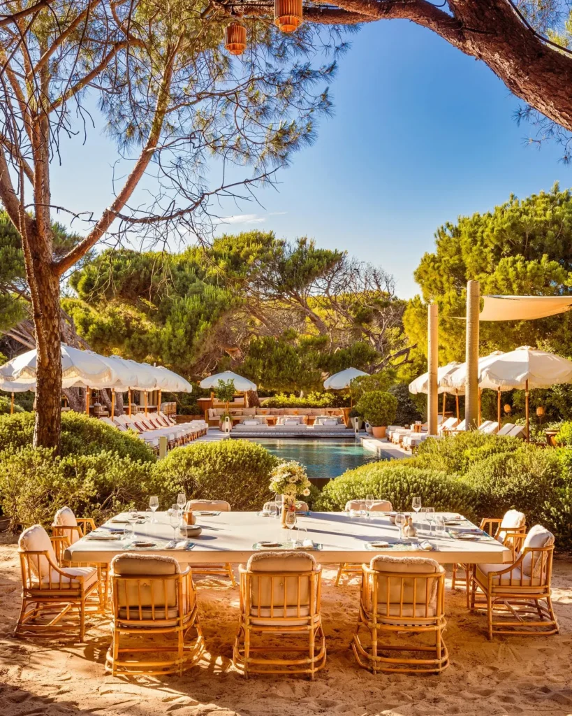 Set dining table on the sand with the pool and parasols beyond at Gigi Rigolatto Saint-Tropez