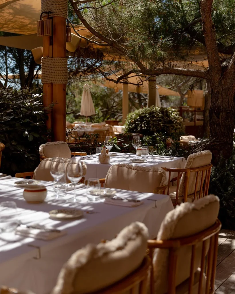 Dappled dining tables with shearling-trimmed rattan chairs among greenery at Gigi Rigolatto Saint-Tropez