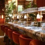 Marble bar lined with rust velvet stools below hanging glasses and greenery at Girafe Paris