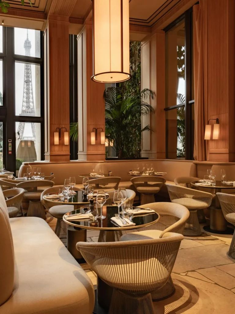 Wire chairs and marble tables by tall windows framing the Eiffel Tower at Girafe Paris