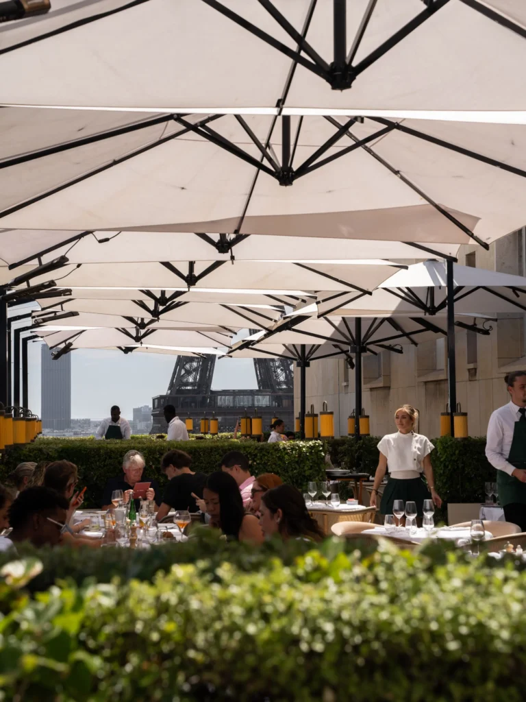 Busy terrace of diners under white parasols with the Eiffel Tower behind at Girafe Paris