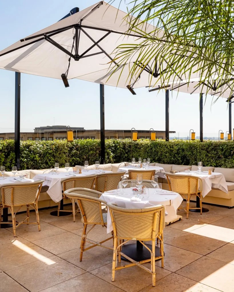 Rattan chairs and white-clothed tables under large parasols behind a hedge at Girafe Paris