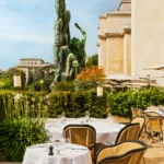 Terrace tables and rattan chairs beside a green bronze statue and the Palais building at Girafe Paris