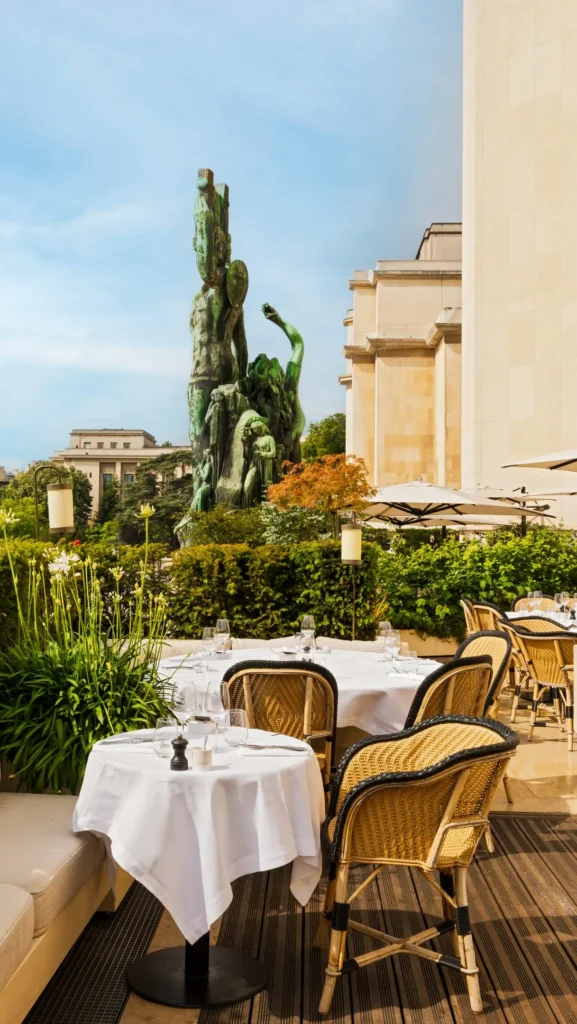 Terrace tables and rattan chairs beside a green bronze statue and the Palais building at Girafe Paris