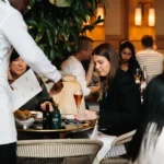 Waiter presenting a menu to a seated guest amid greenery at Girafe Paris