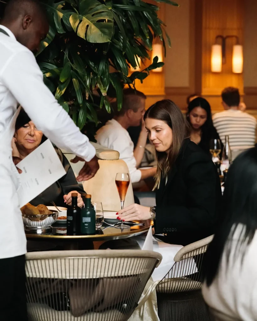 Waiter presenting a menu to a seated guest amid greenery at Girafe Paris