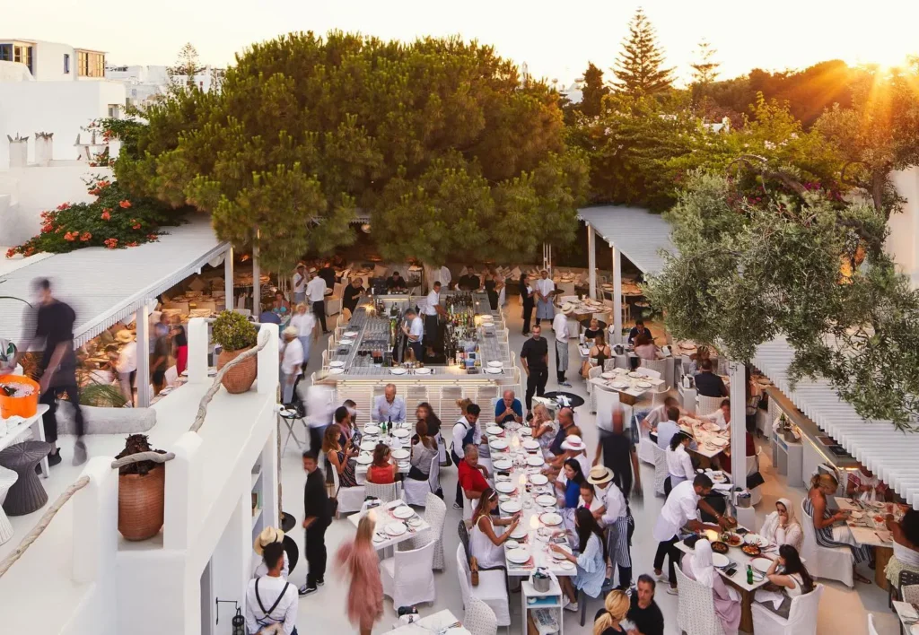 Sunset view over long communal tables and the central bar beneath a large pine at Interni Restaurant Mykonos