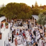 Sunset view over long communal tables and the central bar beneath a large pine at Interni Restaurant Mykonos
