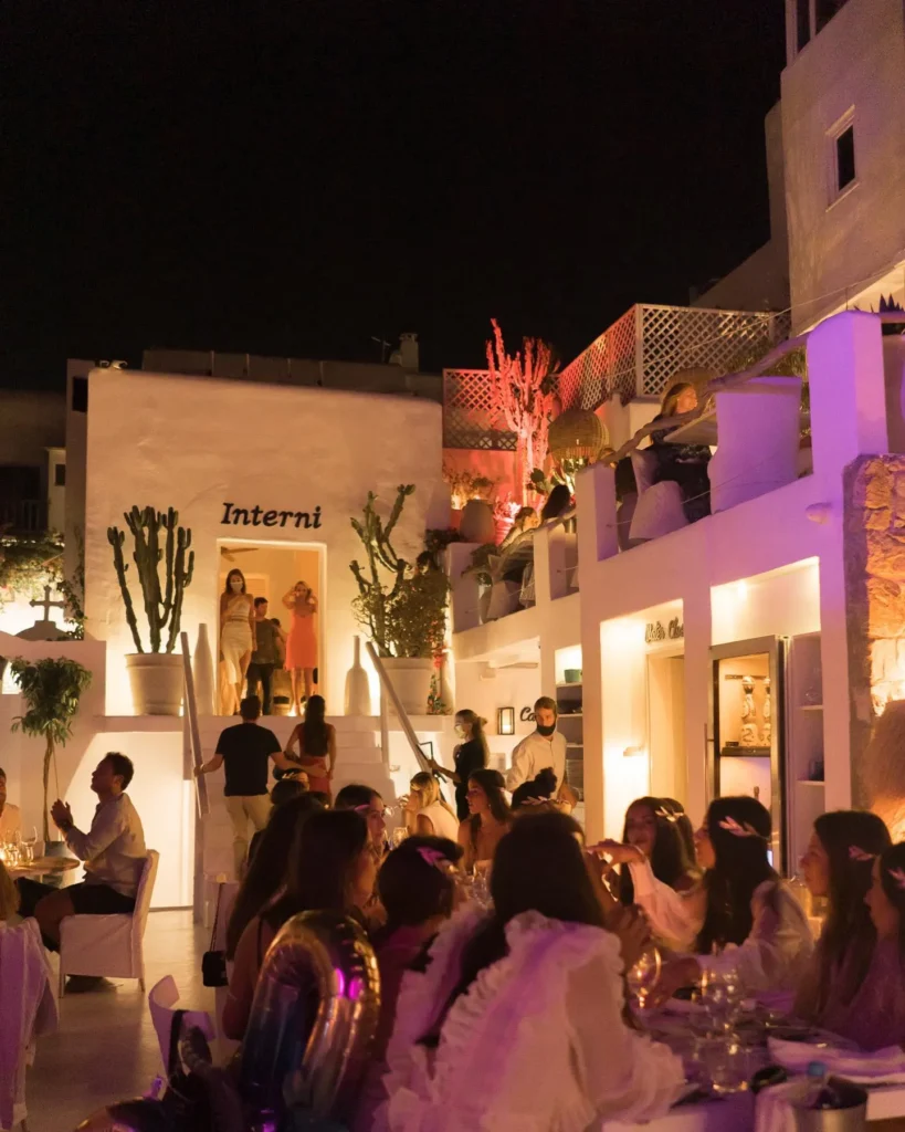 Guests dining beneath the lit Interni sign amid coloured lighting at night at Interni Restaurant Mykonos
