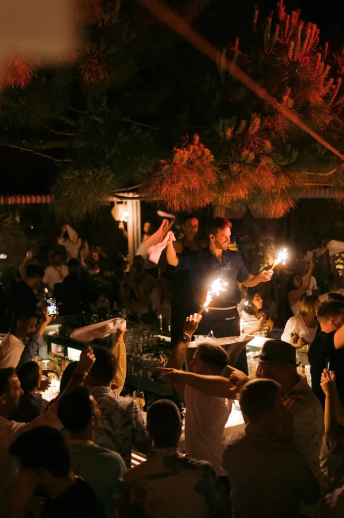 Waiter holding sparklers aloft at the bar amid a cheering crowd at Interni Restaurant Mykonos