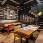 Lower dining area with a live-edge table, coloured chairs and a glass wine cellar beside a spiral staircase at La Bodeguilla Palma