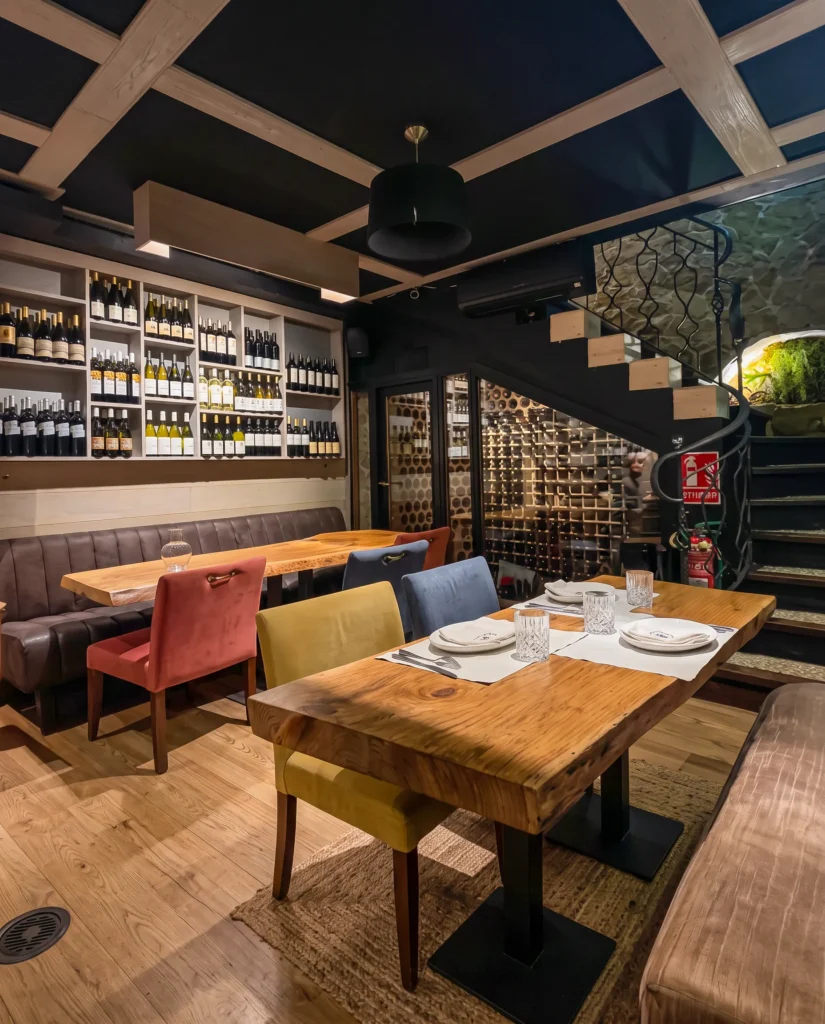 Lower dining area with a live-edge table, coloured chairs and a glass wine cellar beside a spiral staircase at La Bodeguilla Palma