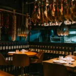 Dining room with tan chairs, brass pendants and rows of hanging jamón and wine at La Bodeguilla Palma