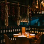 Dining corner beneath hanging jamón, garlic and chilli ristras with a wine-bottle wall at La Bodeguilla Palma