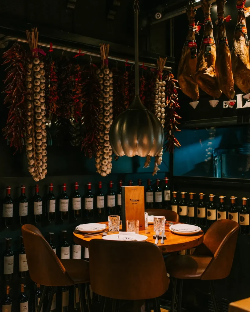 Dining corner beneath hanging jamón, garlic and chilli ristras with a wine-bottle wall at La Bodeguilla Palma
