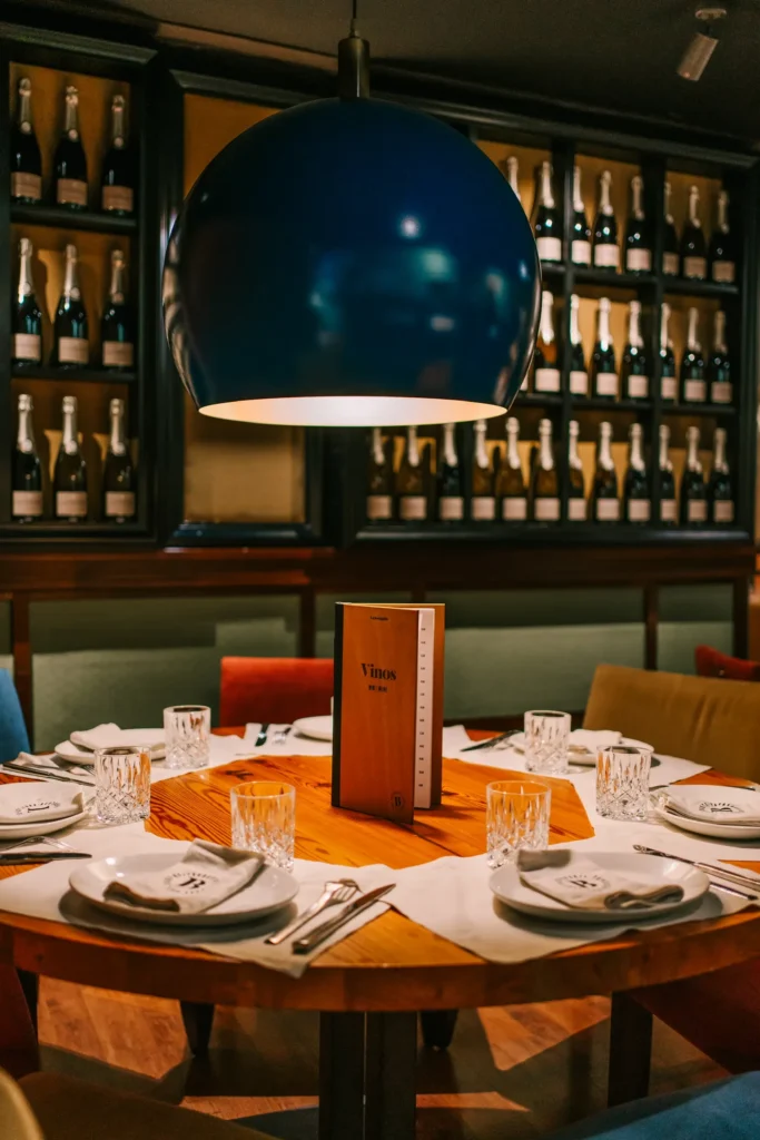 Set round table with a wine list under a large blue globe lamp before a cava-bottle wall at La Bodeguilla Palma