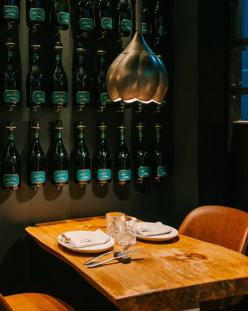 Live-edge table under a brass tulip lamp against a wall of Kripta cava bottles at La Bodeguilla Palma