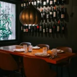 Table with tan chairs under a brass lamp beside a red-wine wall and courtyard window at La Bodeguilla Palma