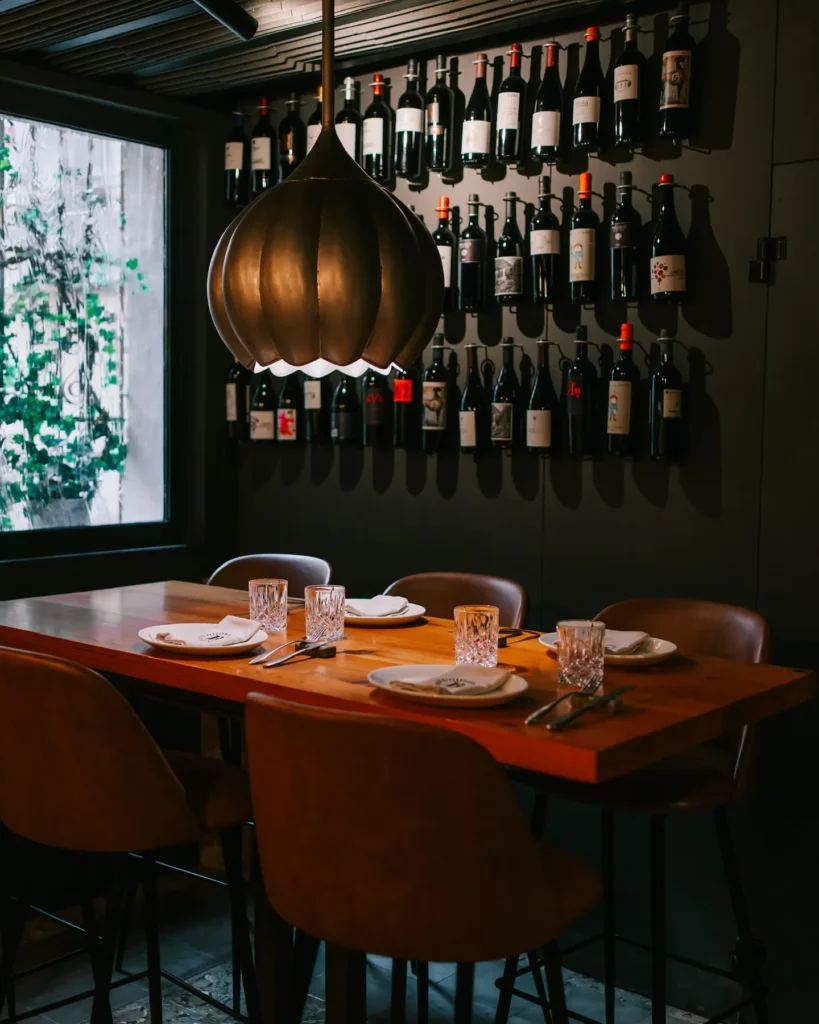 Table with tan chairs under a brass lamp beside a red-wine wall and courtyard window at La Bodeguilla Palma