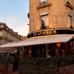 Gold lavenue sign and awning above a fairy-lit hedge in golden evening light at lavenue Paris