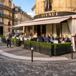 Corner facade with the L'Avenue awning and hedged pavement terrace on a sunny day at L'Avenue Paris