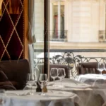 Set tables by a wrought-iron balcony window in daylight beside a studded burgundy wall at lavenue Paris