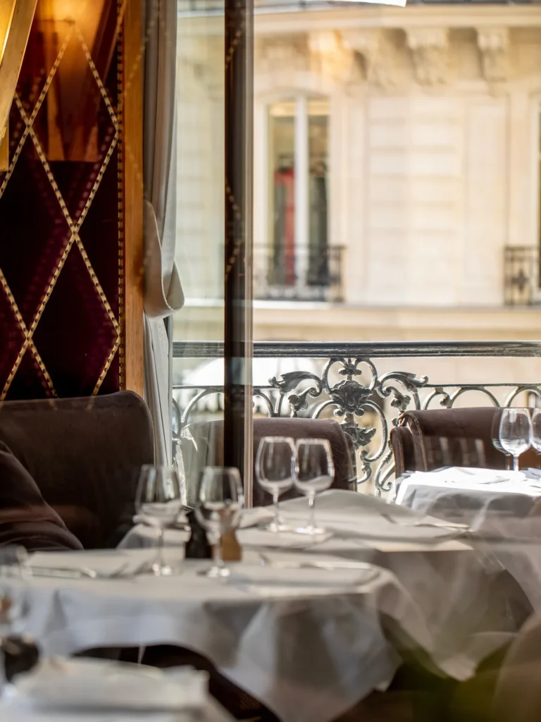 Set tables by a wrought-iron balcony window in daylight beside a studded burgundy wall at lavenue Paris