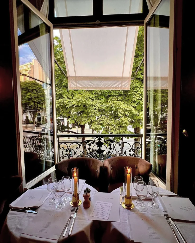 Candlelit table for two set with Avenue menus by an open window over the tree-lined avenue at lavenue Paris