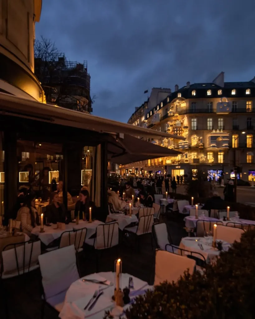 Candlelit terrace tables under the awning with lit Paris buildings beyond at dusk at lavenue Paris