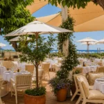 Dining terrace with white parasols, potted trees and sail shades by the sea at Loulou Restaurant Saint-Tropez